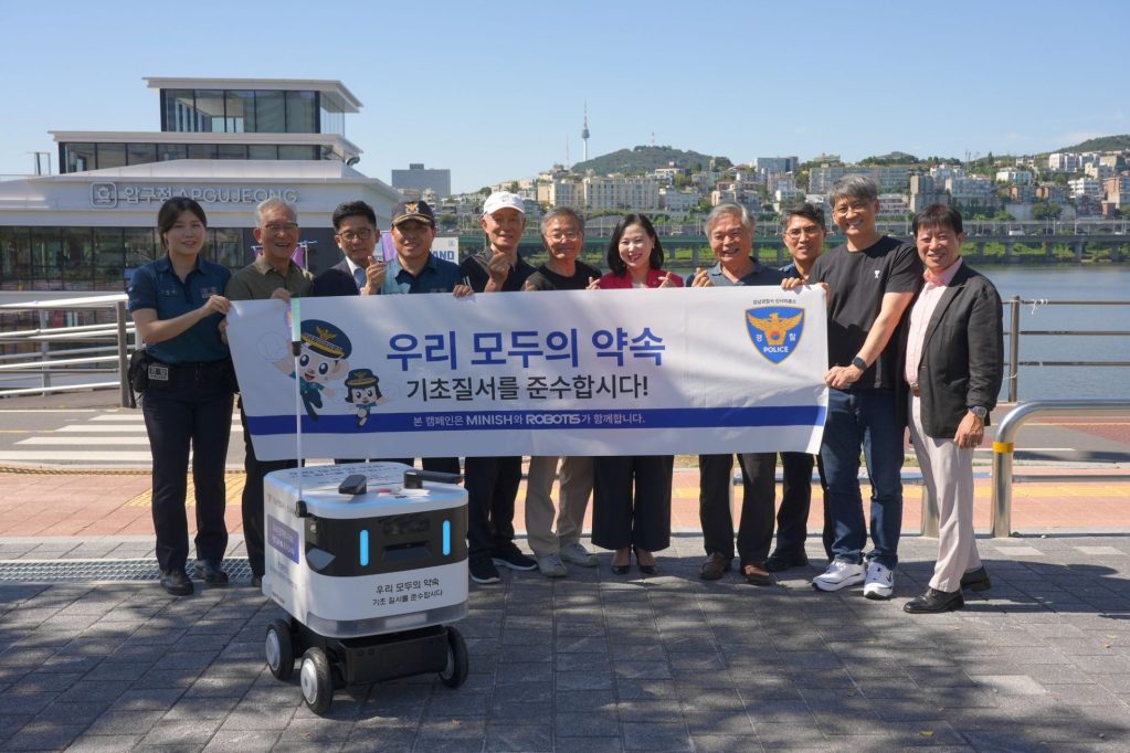 A group of people, including police officers, holding a banner that reads '우리 모두의 약속 기초질서를 준수합시다!' (Let's all promise to maintain basic order!) next to an autonomous delivery robot in a park setting.